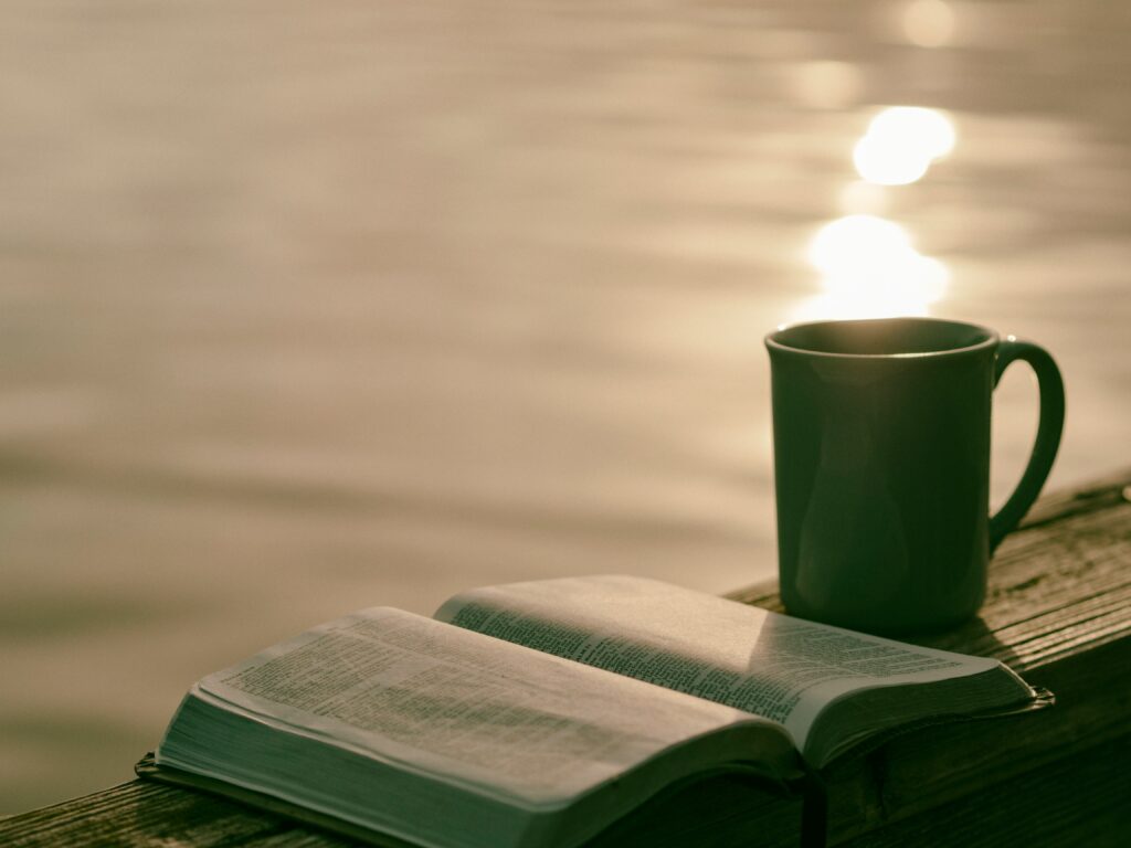 An open book next to a coffee mug on a wooden ledge overlooking the water.