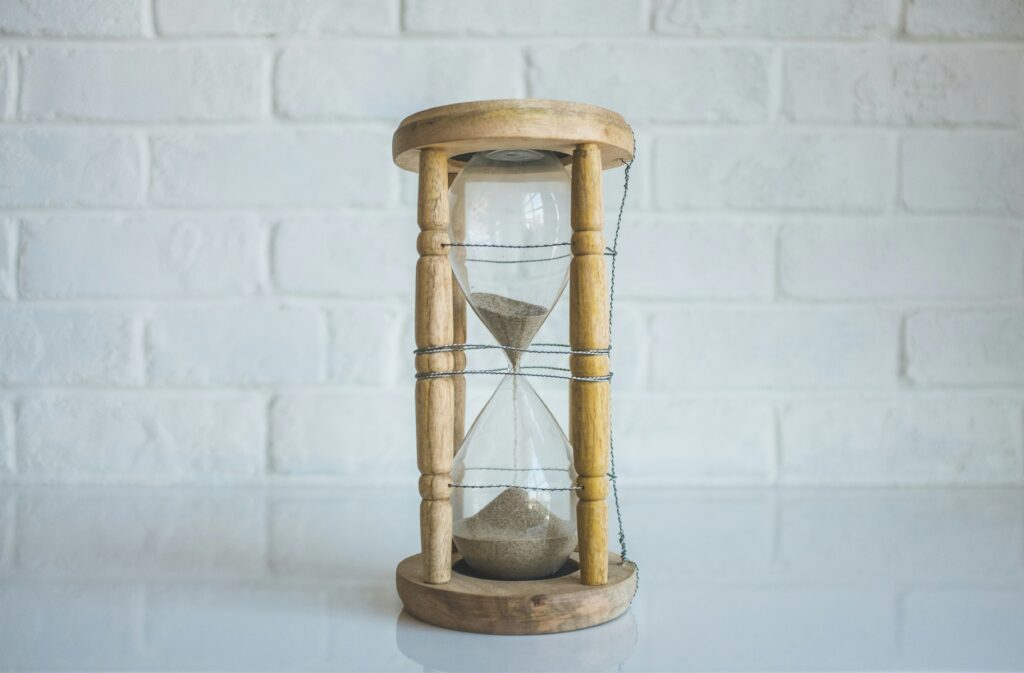 Hourglass with wood frame on a white surface in front of a white brick background.